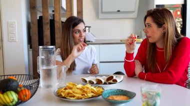 Çağdaş beyaz mutfakta hafta sonu buluşmalarında dinlenirken Meksika aperatiflerinin tadını çıkaran, kahkaha ve nachos paylaşan kadın arkadaşlar.