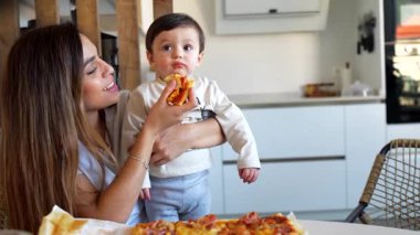 Bakıcı anne, gülümseyen bebeğe pizzayı nazikçe yedirirken aynı zamanda parlak beyaz mutfak ortamında kaynaşıyor.