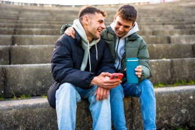 İki adam merdivenlerde oturmuş, yeniden kullanılabilir kahve fincanının tadını çıkarıyor ve birlikte akıllı telefona bakarken gülüyorlar.