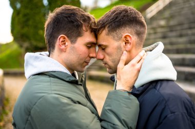Hassas bir anı paylaşan iki adam, alınları birbirine değen, gözleri kapalı, bir park ortamında kucaklaşan