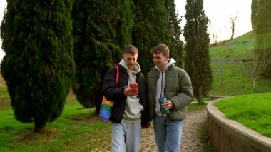 Eşcinsel erkek çift rahat bir park yürüyüşünden zevk alıyor, nazikçe el ele tutuşurken akıllı telefon anlarını paylaşıyorlar, mutluluk ve bağlantı yayıyorlar.