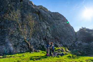 İki kadın dağcı ekipmanlarını, güneşli bir günde zorlu bir tırmanışa hazır, dik bir uçurumun dibinde çimenli bir çayırda hazırlıyorlar.