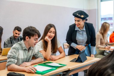 Üniversite öğretmeni sınıftaki iki öğrenciye ders anlatmak için tablet kullanıyor.