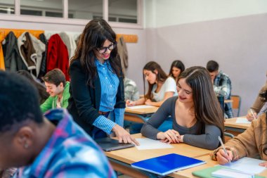 Sınav kağıdıyla gülümseyen bir öğrenciye bir sınıf dolusu öğrencinin sınava girmesi için yardım eden bayan profesör.