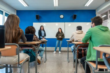 Modern bir üniversite sınıfında sınıf arkadaşlarına ve öğretmenlerine sunum yapan kendinden emin genç bir kadın.