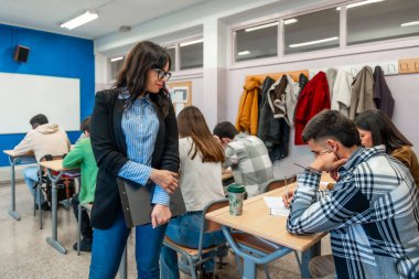 Sınavları bir sınıf ortamında yazan öğrencileri denetleyen, odaklanmış ve akademik bir ortamı teşvik eden üniversite öğretmeni