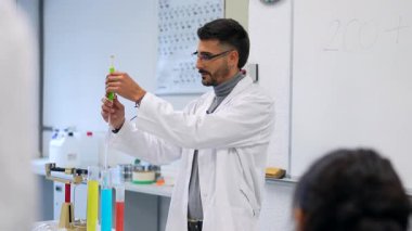 Laboratuvar önlüğü giymiş kimya öğretmeni öğrencilerin önünde laboratuvar malzemelerini kullanarak deney yapıyor.