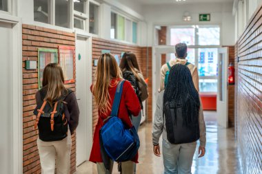 Çeşitli lise öğrencileri kalabalık bir okul koridorunda birlikte yürüyor, sırt çantaları taşıyor ve gülüşmeler paylaşıyor, gençliği ve arkadaşlığı somutlaştırıyorlar.
