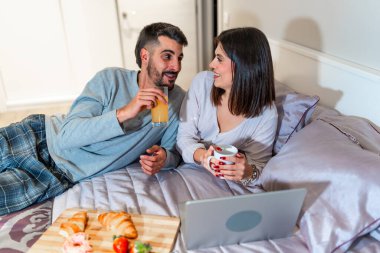 Genç çift yatakta uzanıyor, kahvaltı yapıyor ve laptoplarında bir şeyler izliyor, birlikte kaliteli zaman geçiriyorlar.