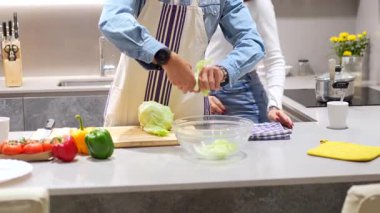 İş birlikçi mutfak sahnesi. Önlüklü bir adam, cam kaseye taze marul yaprakları soyarken partnerin hazırlık sürecini dikkatle izliyor.