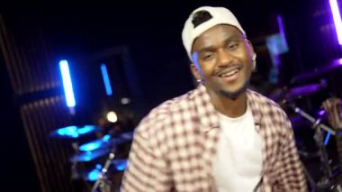 Genç zenci erkek sanatçı, müzik aletleri ve neon ışıklarla çevrili profesyonel müzik stüdyosunda enerjik bir şekilde dans ediyor.