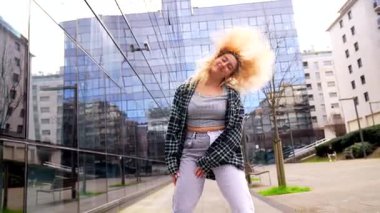 Enerjik genç kadın modern cam binanın önünde özgürce dans ediyor, kent dansı tarzını sergiliyor.