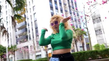 Enerjik genç beyaz kadın canlı bir şehir parkında modern şehir müziğiyle özgürce dans ediyor. Benzersiz tarzını ve hareketlerini sergiliyor.