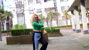 Kendine güvenen genç sarışın modern sokak stili dans ediyor, şehir parkında dinamik bir şekilde hareket ediyor, anlamlı enerji ve ritmik tutku ile.