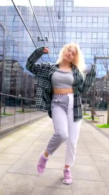 Enerjik genç kadın modern şehrin cam ön planına karşı kendinden emin bir şekilde dinamik kent dansı hareketleri sergiliyor.