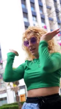 Kıvırcık sarı saçları ve mor güneş gözlükleriyle enerjik genç bir kadın yeşil uzun kollu bluzuyla şehir ortamında özgürce dans ediyor.