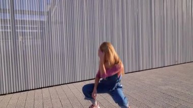 Enerjik genç sarışın kadın modern metalik mimari arka plana karşı şehir tarzı ve sanatsal ifade sergileyerek dinamik sokak dansı hareketleri sergiliyor.