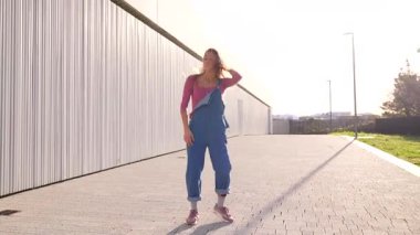 Şehir dans tarzını ifade eden genç sarışın kadın, kot tulumlarıyla dinamik koreografi yapıyor ve şehir kaldırımında pembe bluz giyiyor, yaratıcı hareket ve tutkulu enerji sergiliyor.