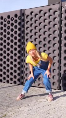 Enerjik sarışın dansçı modern bir mimari arkaplan önünde çeşitli şehir dans figürleri sergiliyor.