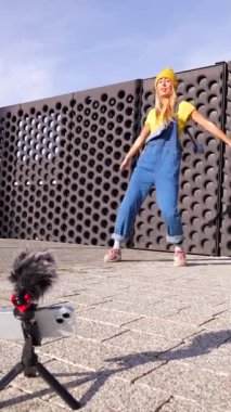 Sarışın genç dansçı dışarıda akıllı telefon ve harici mikrofon kullanarak şehir dansı içeriği yaratıyor.