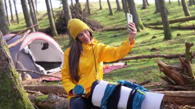 Tek başına kamp yapan genç bir bayan gezgin, parlak sarı yağmurluk giyiyor ve yemyeşil orman çevresinin ortasında çadırın yanında akıllı telefonlardan selfie çekiyor.