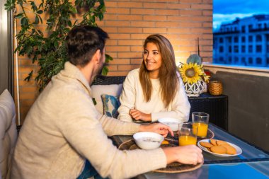 Genç çift, balkonlarında sakin bir akşam yemeği ve sohbet paylaşıyor. Birbirlerinin arkadaşlığından ve şehir manzarasından keyif alıyorlar.