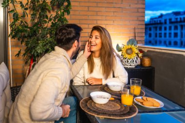 Genç çift gün batımında balkonlarında romantik bir kahvaltı yapıyor. Arkadaşlıklarının ve güzel manzaranın tadını çıkarıyorlar.