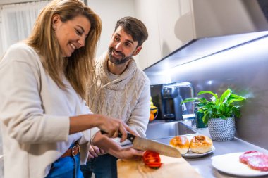Genç çift birlikte yemek hazırlıyor, domates doğruyor ve modern mutfaklarında hamburger yapıyorlar.