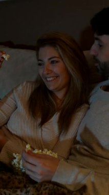 Romantik çift evde film gecesinin tadını çıkarıyor, kaseden patlamış mısır yiyor ve kanepede birlikte gülüyorlar.