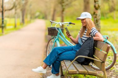 Parktaki bankta yanında bisikletiyle oturmuş güneşli bir günün keyfini çıkarırken akıllı telefon kullanan neşeli genç bir kadın.