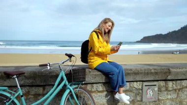 Plaj duvarında dinlenirken yanında bisikletle akıllı telefon kullanan genç bir turist.