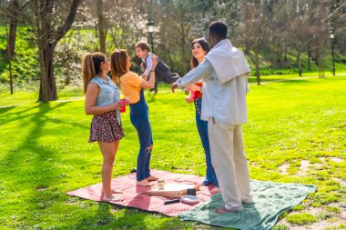 Bir grup neşeli arkadaş parkta güneşli bir günün tadını çıkarıyorlar, piknik sırasında bir çocuğu havaya kaldırıyorlar.