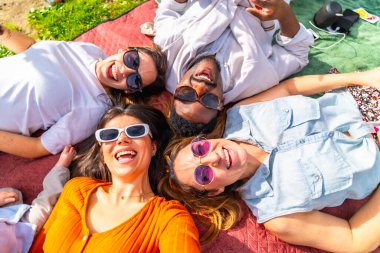 Dört neşeli genç yetişkin birlikte battaniyeye uzanıp güneşli bir parkta pikniğin tadını çıkarıyor, müzik dinliyor ve gülümsüyor.