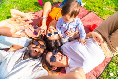 Bir grup neşeli çoklu etnik arkadaş battaniyeye uzanmış güneşli bir piknik sırasında bebekle bir selfie çekiyorlar.