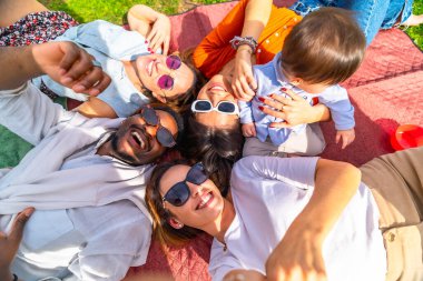 Bir grup neşeli genç ebeveyn parkta güneşli bir piknik gününün tadını çıkarıyor, bebekleriyle selfie çekiyorlar.