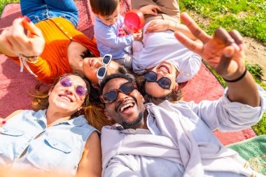 Bir grup neşeli çoklu etnik arkadaş parkta piknik yaparken battaniyenin üzerinde daireler çizerek selfie çekerken boş zamanlarının tadını çıkarıyorlar.