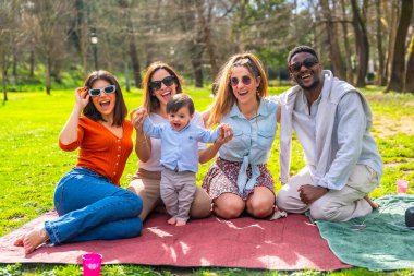Neşeli çoklu etnik arkadaşlar, güneşli bir günde parkta eğlenceli bir pikniğin tadını çıkarıyorlar, bir bebekle gülüp kaynaşıyorlar.