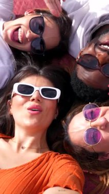 Çeşitli genç arkadaşlar kafa kafaya yatıyorlar, neşe ve dostluk saçıyorlar ve beyaz arka planda şık güneş gözlükleri takıyorlar.