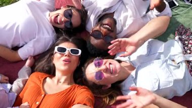Neşeli çoklu etnik arkadaş grubu piknik battaniyesine uzanmış, neşeli selfie ile güneşli park anlarını yakalıyorlar, neşe saçıyor ve kaygısız arkadaşlıklar kuruyorlar.
