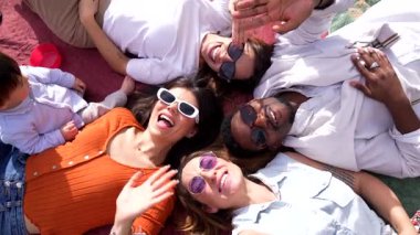 Çoklu etnik aile piknik battaniyesinde uzanır, ebeveynler ve bebekle eğlenceli selfie anları yakalar, güneşli parkın keyfini çıkarırlar.