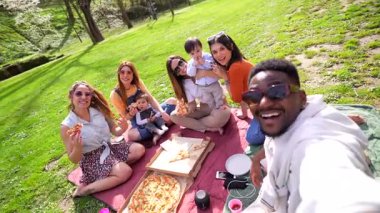 Çoklu etnik arkadaşlar ve çocukları neşeli bir pizza pikniğini paylaşıyor, gülüyor ve aile ve arkadaşlarıyla güneşli park anlarının tadını çıkarıyorlar.