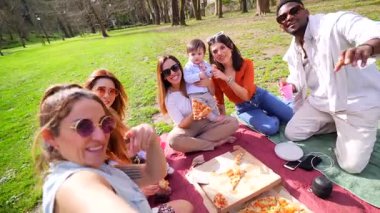 Bir grup arkadaş piknik battaniyesinde dinleniyor. Manzaralı bir parkta bebeklerle yemek yiyip gülüyorlar.