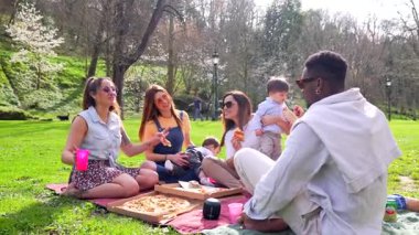 Neşeli bir grup arkadaş pizza ve içki paylaşıyor, güneşli bir günde halka açık bir parkta arkadaşlığı kutluyorlar.