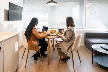 İki iş kadını kahvaltıdan zevk alıyor ve iş gezileri sırasında modern bir otel odasında dizüstü bilgisayar ve tabletle çalışıyorlar.