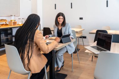 İki iş kadını kahvaltıdan zevk alıyor ve iş gezileri sırasında bir tablet üzerinde çalışıyorlar, otel ortamında iş ve eğlence karışımı sergiliyorlar.