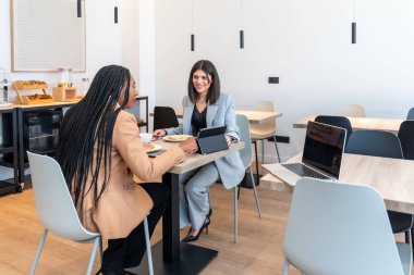 İki iş kadını kahvaltı yapıyorlar ve iş gezileri sırasında bir otel restoranında dijital tablet ve dizüstü bilgisayarla çalışıyorlar.