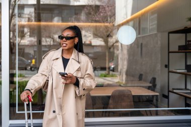 Şık bir iş kadını, bir sonraki iş seyahatine hazır bir şekilde, bir otel lobisinin önünde el çantasıyla beklerken akıllı telefonunu kullanıyor.