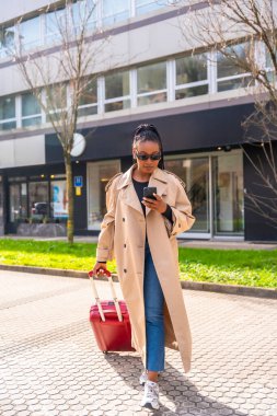 Şık bir iş kadını bavulu çekiyor ve iş gezisinde otelin dışında yürürken akıllı telefon kullanıyor.