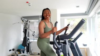 Siyah kadın yönetici, eliptik makine üzerinde egzersiz yapıyor, iş gezisi sırasında sağlık rutinini sürdürüyor, otel jimnastik salonunda doğal güneş ışığı penceresinden içeri giriyor.