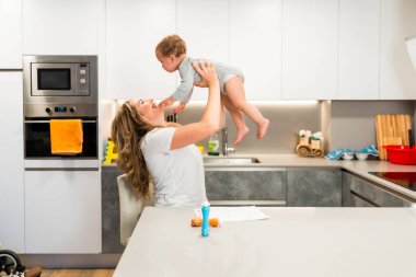 Mutlu anne bebekle oynuyor, modern bir mutfakta onu havaya kaldırıyor, birlikte vakit geçiriyorlar.